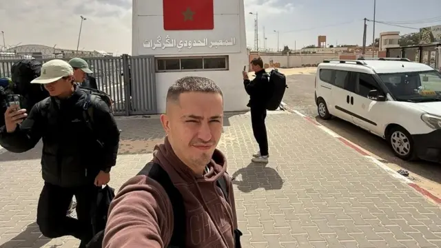 Tom Ruck prend un selfie devant le poste frontière de Guerguerat entre le Sahara occidental et la Mauritanie. Derrière lui se trouve une grande structure en béton sur laquelle est peint un drapeau marocain.